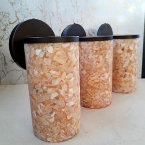 May include: Three cylindrical wall sconces with a mosaic design. The sconces have a black metal base and a cylindrical shade filled with light-colored, irregularly shaped pieces. The shades are topped with a black metal cap.