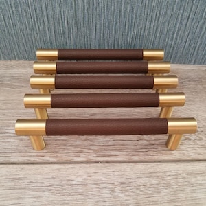 May include: Set of five cabinet pulls. Each pull features a cylindrical, textured brown leather grip with gold-toned metal ends and a T-shaped base. The hardware is arranged on a light wood surface.