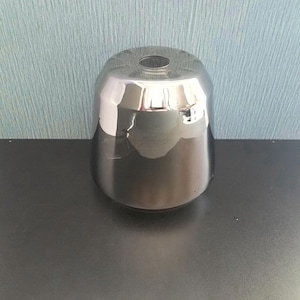 May include: A shiny, silver-colored metal lampshade with a flared, conical shape. The lampshade has a smooth, reflective surface and a small opening at the top. The design is modern and minimalist.