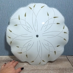 May include: A white, scalloped-edge ceiling light fixture with a floral design. The fixture has a central black dot and is decorated with thin, curved lines and gold-colored leaf-like shapes. The light is on a wooden surface.