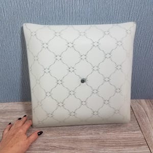 May include: Square, white light fixture with a gray geometric pattern. The fixture has a central, dark-colored button and is set against a light blue wall and wooden surface.