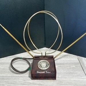 May include: Vintage Directronic Color TV antenna. The antenna features a dark brown base with the text "Directronic Color". It has two gold-colored, adjustable arms and two silver-colored circular hoops. A black cord is attached to the base.