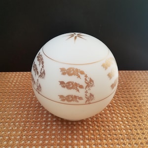 May include: A white, spherical decorative container with a lid. The container is adorned with gold floral patterns and a gold starburst design on top. A gold band encircles the middle. The container sits on a light brown surface.