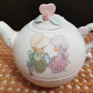 May include: White ceramic teapot with a lid featuring a pink heart and rose accent. The teapot is decorated with a graphic of two girls pouring tea. The spout and handle have floral details. A charming collectible.