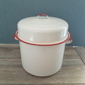 May include: A cylindrical white enamel stockpot with a matching lid. The pot features a thin, vibrant red rim around the top edge and two small side handles. The lid has a small, arched handle on top, and the entire exterior is smooth and glossy.