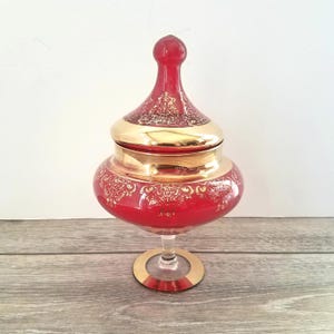 May include: A red glass jar with a gold-colored lid and base. The jar has a decorative floral pattern in gold. The jar is on a clear glass stem with a gold base. The jar is likely used for storage or decoration.