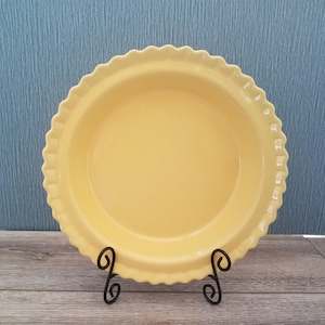 May include: A round, yellow ceramic pie dish with a decorative scalloped edge. The dish is displayed upright on a black metal wire stand, featuring a smooth, glossy finish and a wide, shallow basin designed for baking various types of pies.