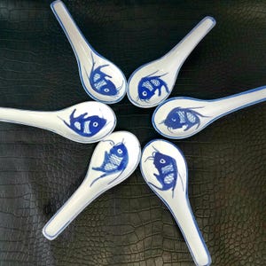 May include: Six white ceramic soup spoons with a blue fish design. Each spoon has a blue rim and a blue fish illustration in the bowl. The spoons are arranged in a circular pattern.