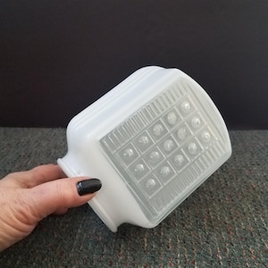 May include: A white, rectangular glass light fixture shade with a textured, grid-like pattern on the front. The shade has a rounded top and a flared base. The shade is held by a hand with dark nail polish.