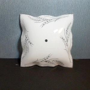 May include: A square, white glass light fixture with a wavy edge. The fixture is decorated with black wheat stalk designs. A small, round, black detail is in the center.