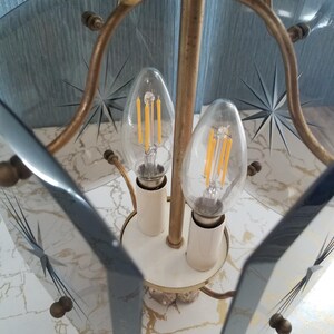 May include: A vintage brass and glass chandelier with two clear light bulbs. The glass panels have starburst designs etched into them. The fixture has a white base and is set against a marble-patterned surface.