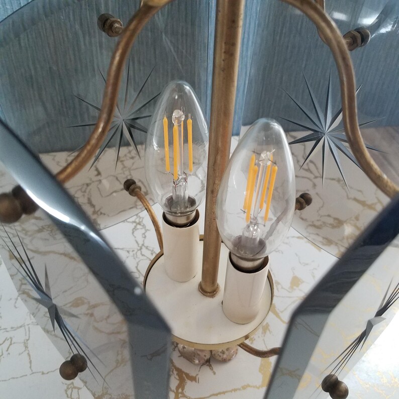 May include: A vintage brass and glass chandelier with two clear light bulbs. The glass panels have starburst designs etched into them. The fixture has a white base and is set against a marble-patterned surface.