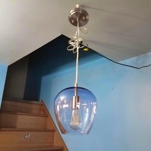 May include: A blue glass pendant light fixture with a brushed nickel finish. The light has a teardrop-shaped glass shade and a visible Edison-style bulb. The fixture is suspended from the ceiling by a white cord and a brushed nickel canopy.