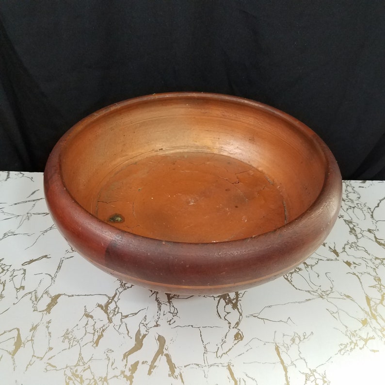May include: A round wooden bowl with a reddish-brown exterior and a lighter brown interior. The bowl has a wide rim and sits on a white surface with gold veining. The background is black.