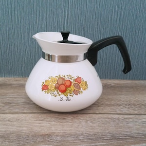 May include: A white teapot with a black handle and lid, featuring a silver metal band around the neck. The body displays a colorful botanical illustration of fruits and flowers with the text Le Thé printed in black script below the design.