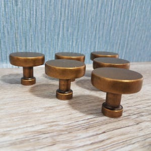 May include: Six bronze-colored cabinet knobs with a round top and a cylindrical base. The knobs are arranged on a light wood surface, with a blue textured background. These knobs are suitable for furniture restoration or new builds.