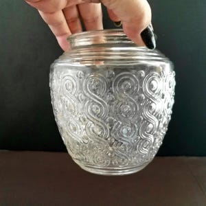 May include: A clear glass jar with a decorative swirl pattern. The jar has a wide mouth and a rounded body. The jar is held by a hand with a black painted fingernail. The background is dark.