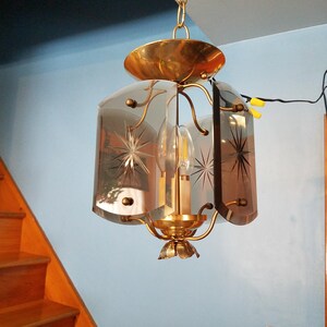 May include: Vintage brass pendant light fixture with etched starburst designs on the glass panels. The light has a decorative floral base and a gold-toned canopy. The fixture is suspended from a chain.
