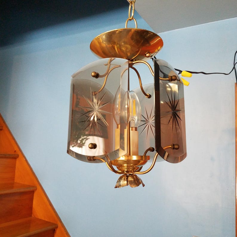 May include: Vintage brass pendant light fixture with etched starburst designs on the glass panels. The light has a decorative floral base and a gold-toned canopy. The fixture is suspended from a chain.