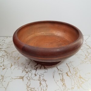 May include: A round, wooden bowl with a wide rim and a pedestal base. The bowl is a deep reddish-brown color, with a lighter, orange-brown interior. The bowl sits on a white surface with gold veining.
