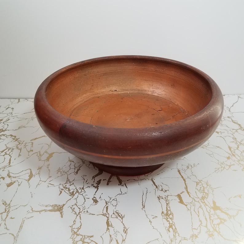 May include: A round, wooden bowl with a wide rim and a pedestal base. The bowl is a deep reddish-brown color, with a lighter, orange-brown interior. The bowl sits on a white surface with gold veining.