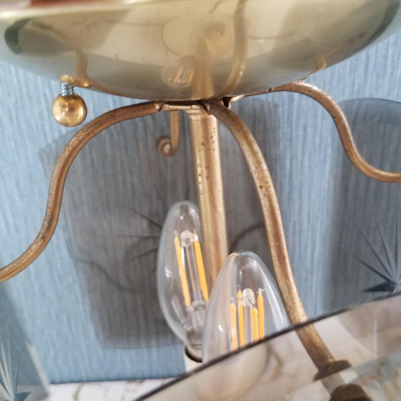 May include: Close-up of a gold-colored chandelier with two clear light bulbs. The chandelier has a bowl-shaped top and curved arms. The light bulbs have a yellow filament. The background is a light blue color.