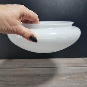 May include: A white, opaque glass light fixture shade with a rounded bowl shape. The shade has a slightly flared rim and a smooth, glossy surface. The shade is held by a hand with a black painted fingernail.
