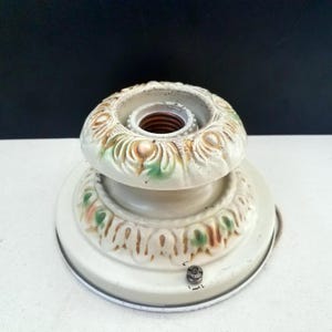 May include: Off-white ceramic light fixture with decorative floral accents in green, pink, and brown. The fixture has a circular base and a bulb socket. The design is vintage and ornate.