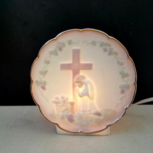 May include: A round, off-white porcelain night light with a scalloped edge and gold trim. The design features a pink cross, a kneeling figure, and grape vines. The light illuminates the scene from within.