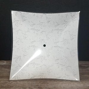 May include: A square glass light shade featuring a white background with a repeating light gray swirling floral pattern and a single circular hole centered in the middle of the piece for mounting purposes.