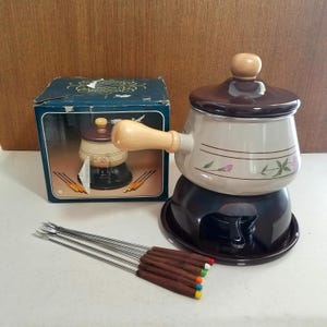 May include: A vintage fondue set with a dark brown base, a cream-colored pot with floral accents, and a matching lid. The set includes a box and several fondue forks with wooden handles and colorful tips.