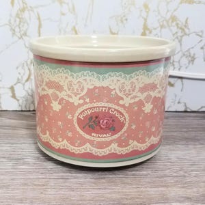 May include: A cylindrical ceramic potpourri crock with a cream-colored rim and base. The exterior features a pink background with a repeating pattern of small flowers, overlaid with a cream-colored lace design and a central oval label reading Potpourri Crock RIVAL with a rose illustration.