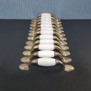 May include: A collection of cabinet handles with a brass finish and white ceramic center. The handles are arranged in rows, showcasing their design. The hardware is suitable for furniture restoration or new builds.