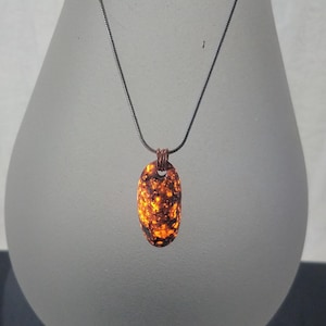 May include: A pendant necklace featuring an oval-shaped stone with a speckled orange and brown pattern. The stone is suspended from a thin, dark silver chain. The pendant is attached to the chain with a copper-colored loop.