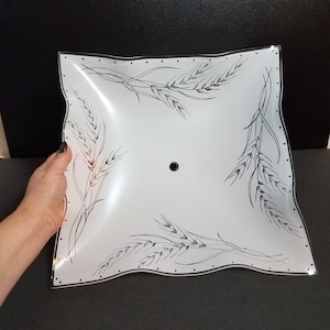 May include: A square, white light fixture with a wavy edge and black wheat stalk design. The fixture has a black center hole and is held by a hand. The wheat stalks are in each corner.