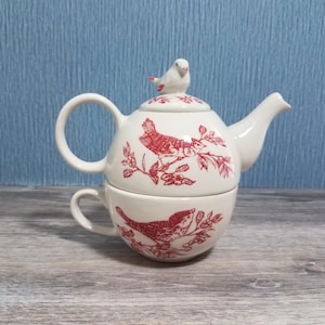 May include: A cream-colored ceramic tea set for one, consisting of a teapot stacked on a teacup. Both pieces feature a red botanical print of birds perched on branches. The teapot lid is topped with a small, sculpted white bird figurine.