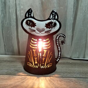 Puede incluir: Una linterna de Halloween con forma de gato de metal negro con una cara blanca y un diseño de esqueleto. La cara del gato tiene patrones decorativos, y el cuerpo presenta un esqueleto brillante. La cola tiene una araña al final.
