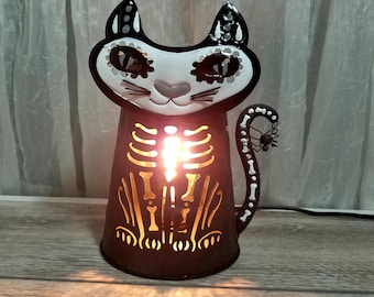Może przedstawiać: Czarna, metalowa latarnia na Halloween w kształcie kota z białą twarzą i wzorem szkieletu. Twarz kota ma dekoracyjne wzory, a ciało świecący szkielet. Ogon ma pająka na końcu.