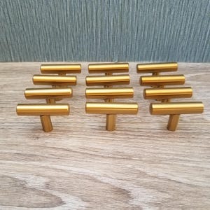 May include: A collection of ten gold-colored T-bar cabinet pulls arranged on a wooden surface. Each pull has a cylindrical bar and a cylindrical base. The pulls are evenly spaced and appear to be new.