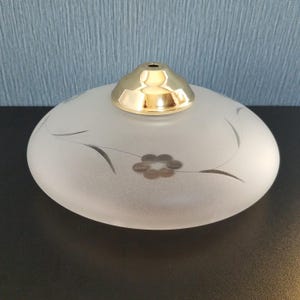 May include: Frosted glass lampshade with a gold-colored metal cap. The lampshade has a floral design with a flower and curved lines. The lampshade is oval-shaped and sits on a dark surface.