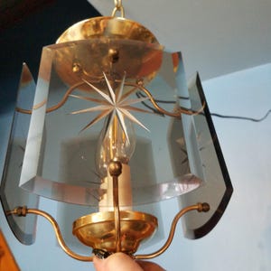 May include: Vintage pendant light fixture with a starburst design etched into the clear glass panels. The fixture has a brass-colored metal frame and a single light bulb. The glass panels are geometric and angled, creating a unique lighting effect.