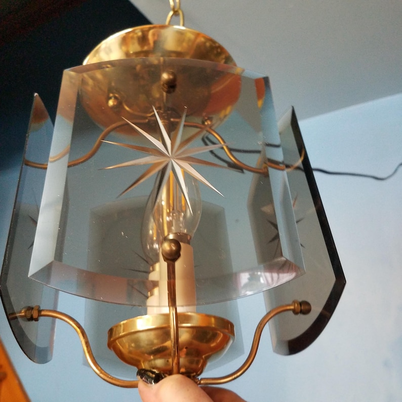 May include: Vintage pendant light fixture with a starburst design etched into the clear glass panels. The fixture has a brass-colored metal frame and a single light bulb. The glass panels are geometric and angled, creating a unique lighting effect.