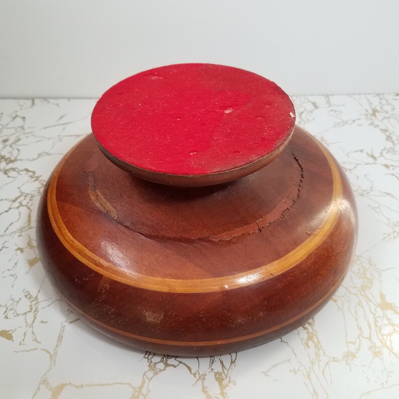 May include: A wooden pedestal bowl with a red felt-covered top. The bowl is made of dark brown wood with a lighter brown and tan stripe around the middle. The bowl has a round base and a slightly cracked surface.
