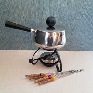 May include: A vintage fondue set with a stainless steel pot, black handle, and lid with a black knob. The set includes a black metal stand and several fondue forks with wooden handles and colorful tips.