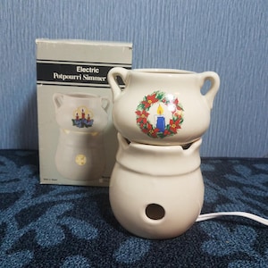 May include: An off-white ceramic electric potpourri simmer with two handles and a Christmas wreath graphic featuring a blue candle. The device sits in front of its original packaging box, which displays the text Electric Potpourri Simmer.