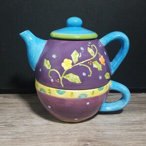 May include: A ceramic tea-for-one set featuring a purple teapot with a blue lid and handle, decorated with yellow and orange flowers, green leaves, and white dots. The base is a purple cup with a yellow band featuring green and blue circular patterns.