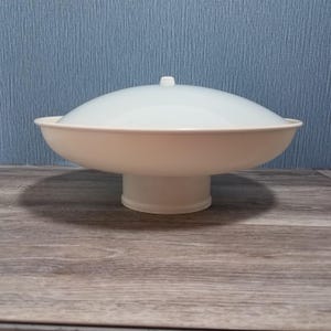 May include: A white pedestal bowl featuring a wide, shallow basin and a matching translucent, frosted white dome lid with a small central knob. The structure is supported by a cylindrical base, creating a balanced, elevated serving dish design.