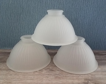 May include: Three bell-shaped lamp shades made of frosted glass with a vertical ribbed texture. Each shade features a circular opening at the top for mounting and a wide, flared base. The glass has a semi-translucent, matte white finish.
