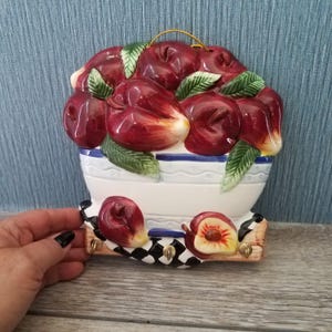 May include: A ceramic wall hanging featuring a white bowl with a blue stripe, overflowing with glossy red peaches and green leaves. The base has a black and white checkered pattern and three gold hooks for keys.