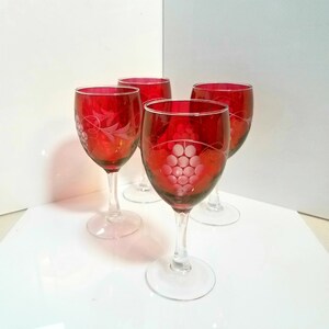 May include: Four ruby red wine glasses with clear stems and bases. Each glass features an etched grape design. The glasses are arranged on a white surface, showcasing their elegant design and color.